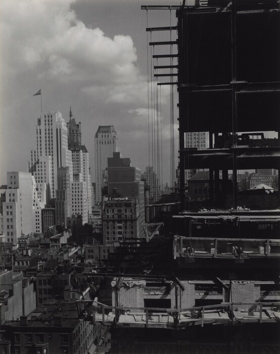 The image shows an urban scene with completed buildings and a partially constructed high-rise. A tall building under construction with steel beams is visible on the right. Completed skyscrapers are seen on the horizon. Different architectural styles are present, from art deco to modern buildings. The sky has clouds, adding depth. The black and white color scheme emphasizes light and shadow contrasts, creating a dramatic effect. It captures a moment of urban development with finished and ongoing construction.