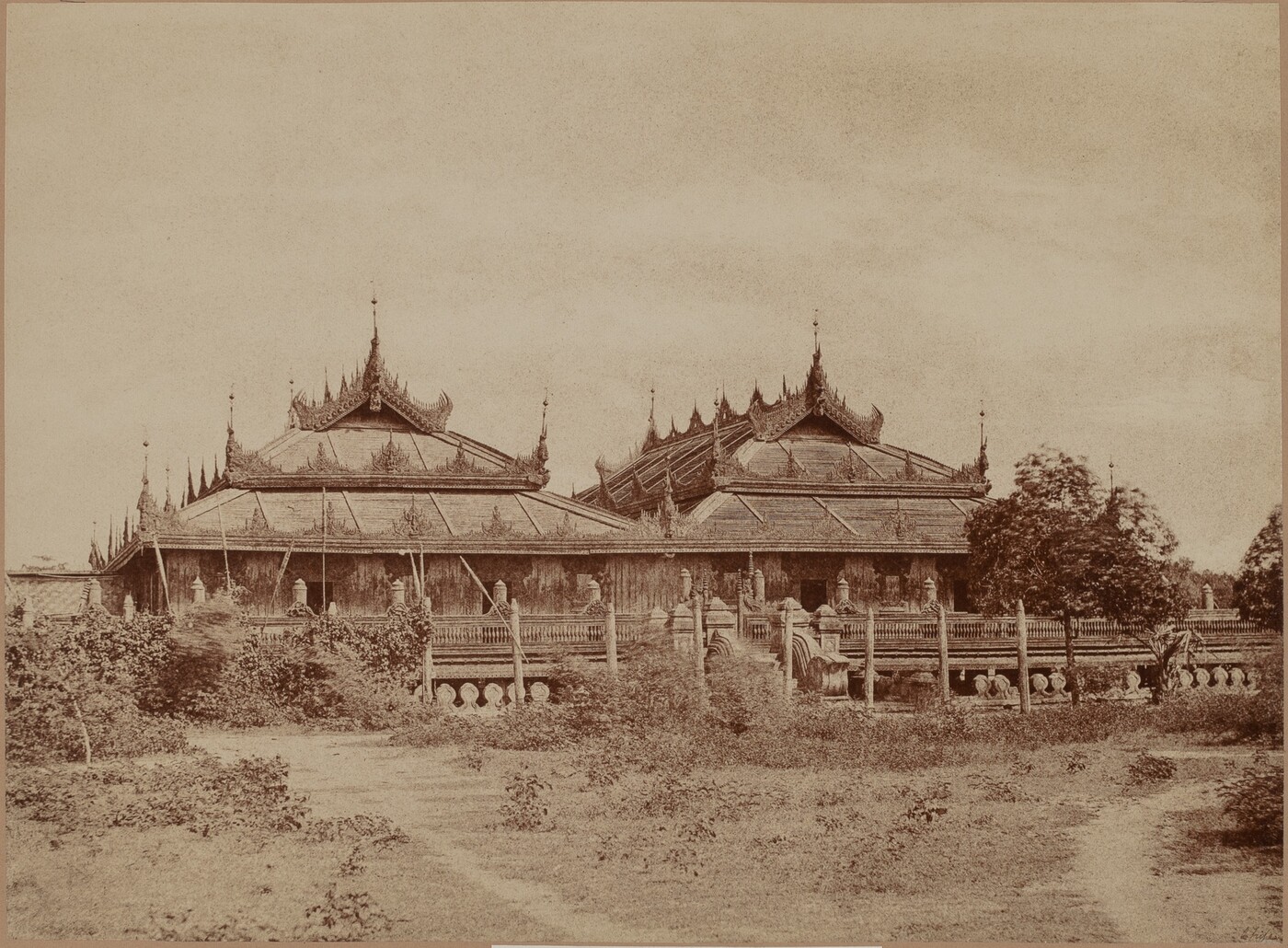 The image shows a traditional building with intricate architecture, pointed spires, and ornate detailing. The perspective is slightly elevated, providing a comprehensive view of the structure. The rooflines are prominent, leading the viewer's gaze upward. The color palette is monochromatic with sepia tones. Vegetation and pathways lead up to the building in the foreground, surrounded by a low fence and shrubbery. Trees are visible in the distance. The sky above is clear.
