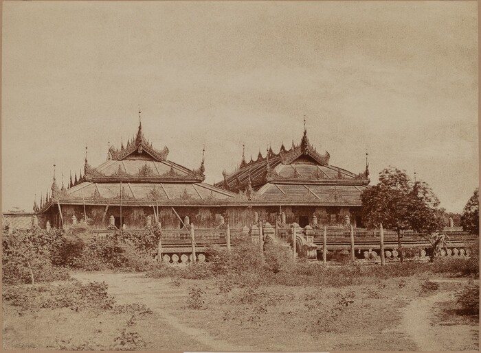The image shows a traditional building with intricate architecture, pointed spires, and ornate detailing. The perspective is slightly elevated, providing a comprehensive view of the structure. The rooflines are prominent, leading the viewer's gaze upward. The color palette is monochromatic with sepia tones. Vegetation and pathways lead up to the building in the foreground, surrounded by a low fence and shrubbery. Trees are visible in the distance. The sky above is clear.