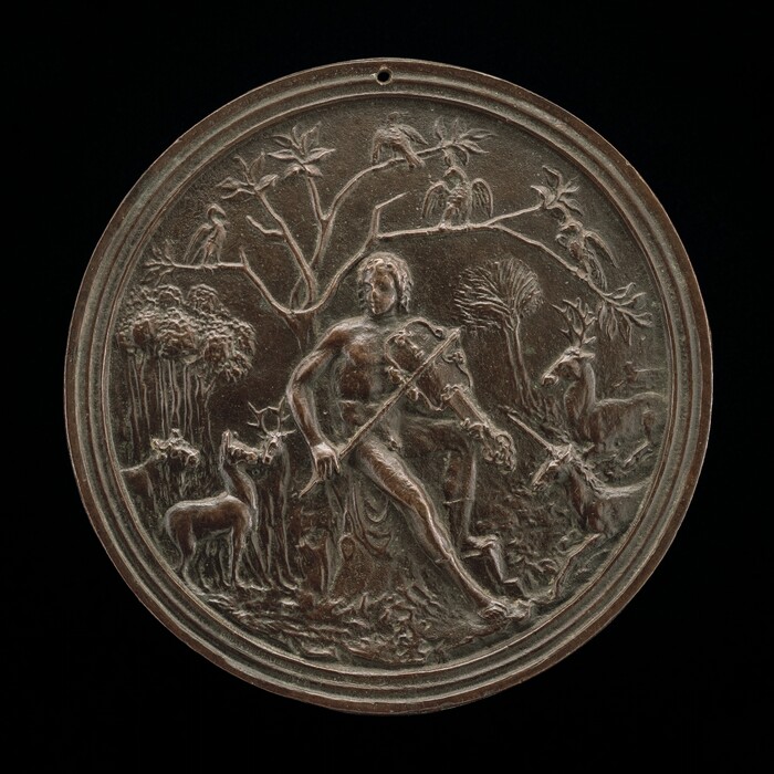 This round relief depicts a man leaning against a tree, playing a lyre or violin with a long bow. Surrounding him are various animals, including several deer, a unicorn, and birds that rest on the tree branches above him. The man is centered in the forest setting, looking to the left, with trees and foliage in the background. The sculpture is made of a dark brown material, and there is a small hole on the top of it.