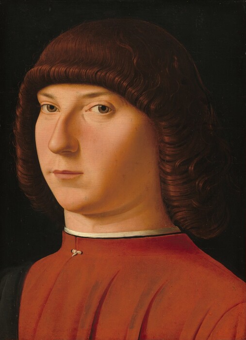 Shown from the chest up, a clean shaven, young man with pale, rosy skin and curly, mahogany-red hair, looks at or toward us against a black background in this vertical portrait painting. His body is angled to our left, and he turns his face slightly to look our way with sienna-brown eyes under curved eyebrows. He has a long, prominent nose and small thin pink lips. His shoulder length hair curls tightly around his face and covers his ears. He wears a tomato-red shirt with a high collar trimmed with white.