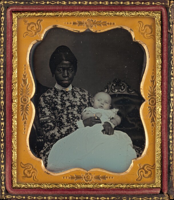 The image shows a seated woman visible from the waist up, holding a baby in her lap. She is positioned straightforward, facing the camera. The woman has dark skin, a defined nose, and styled back hair. Her clothing is a patterned dress with a high collar and a small embellishment. The baby is dressed in a light-colored gown. The woman wears a small necklace or pendant around her neck. The background includes a decorative chair, and the photograph is framed in an elegant, gold-patterned frame.