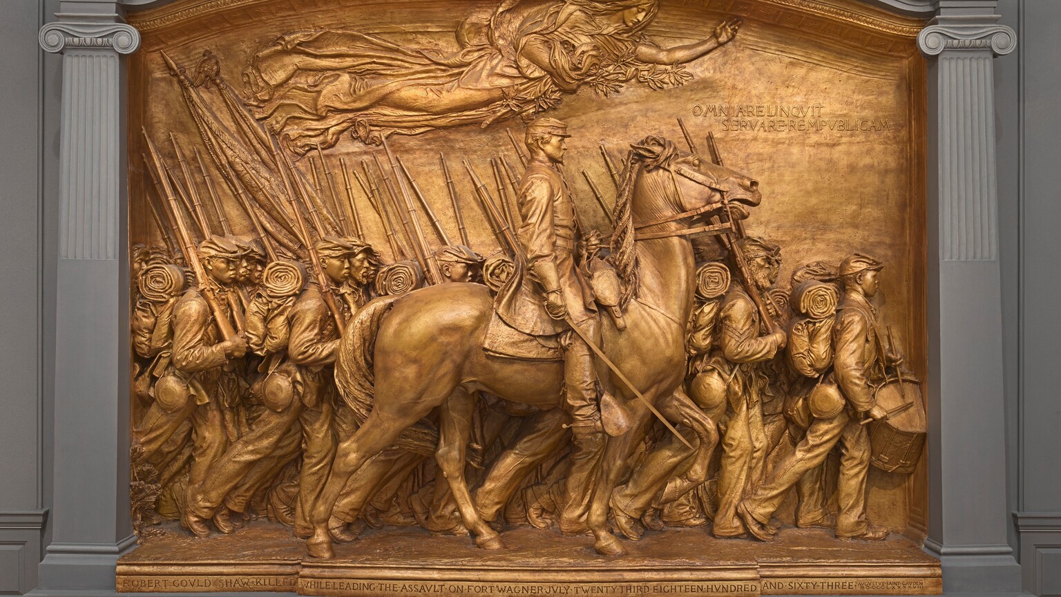 A white man in military uniform rides a horse in front of a regiment of five rows of Black troops in this sculpture, which is painted entirely in gold. The artist created a shallow, stage-like space with an arched top so the men are sculpted in three dimensions, though they become more compressed as they move back in space. The men and horse face our right in profile in this view. The man on the horse has a pointed, straight nose and a goatee. He wears a cap with a flat top and narrow brim, a knee-length coat, gloves, and knee-high boots with spurs. He holds a thin sword down by the side of the horse with his right hand and holds the reins of the horse with his left. The horse’s head is pulled upward by the short reins, and its mouth is open around the bit. About twenty soldiers are lined up in rows beyond the horse, and they march in unison. They carry blankets rolled atop knapsacks, canteens, and rifles resting on their right shoulders. However, the details of how their uniforms bunch up around their equipment and the way their caps have been molded and fit is unique to each person. Their ages also vary from young and cleanshaven to bearded, older men. Two men carry furled flags near the back, to our left, and a drummer boy plays at the head of the regiment, to our right. All the men look straight ahead, their lips closed. A woman in a billowing robe floats above them under the arched top of the sculpture with her eyes closed. Her left arm is outstretched, and she holds a laurel branch and poppies close to her body with her right arm. An inscription in the upper right corner is created with raised capital letters: “OMNIA RELINQVIT SERVARE REMPVBLICAM.” A longer inscription is carved into the base along the bottom edge of the memorial, also in all caps: “ROBERT GOVLD SHAW KILLED WHILE LEADING THE ASSVLT ON FORT WAGNER JVLY TWENTY THIRD EIGHTEEN HVNDRED AND SIXTY THREE.” The artist’s signature is inscribed In the lower right corner, in smaller letters: “AVGVTVS SAINT GAVDEN M-D-C-C-C-L X X X X V I I I.”