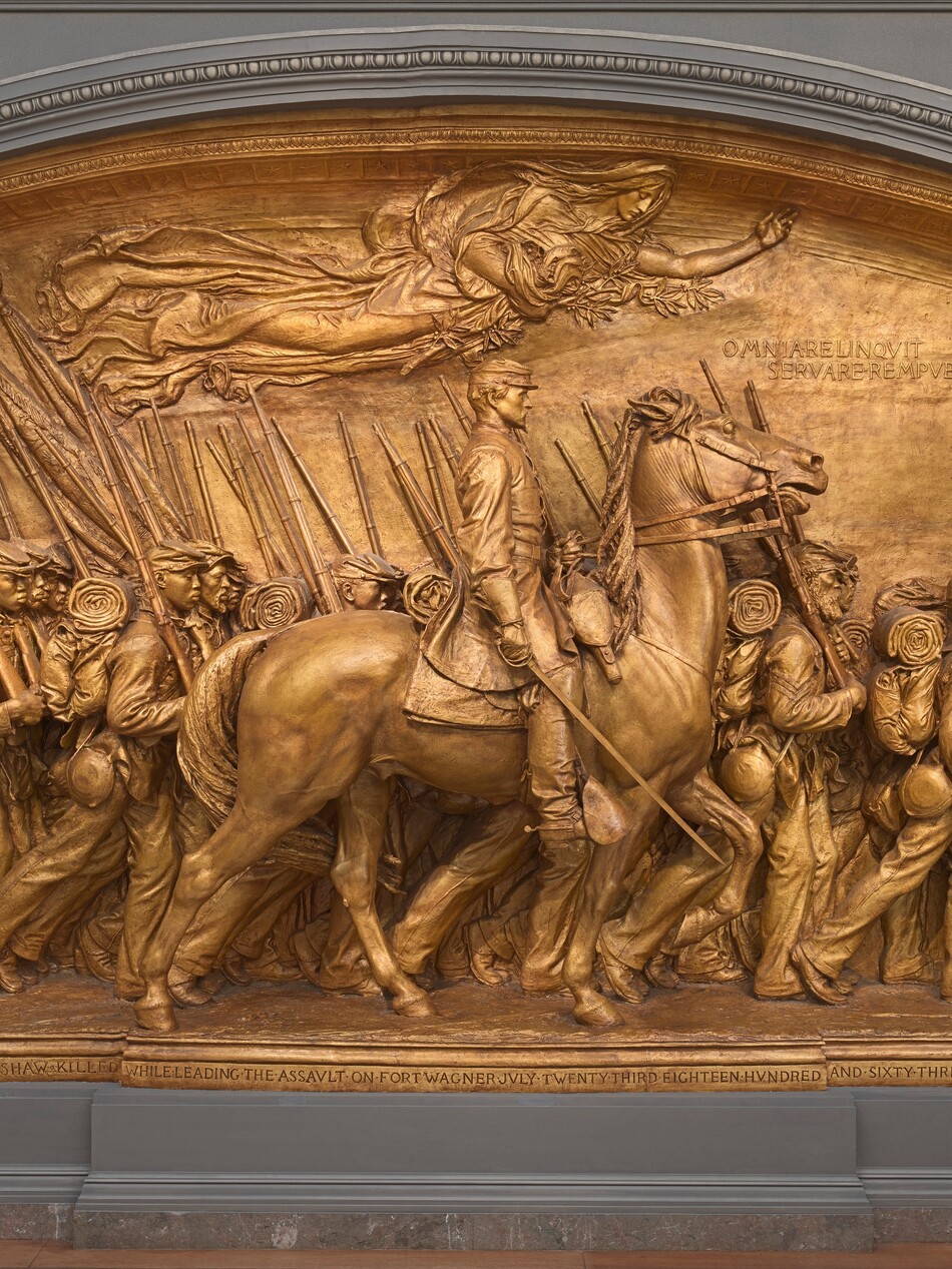 A white man in military uniform rides a horse in front of a regiment of five rows of Black troops in this sculpture, which is painted entirely in gold. The artist created a shallow, stage-like space with an arched top so the men are sculpted in three dimensions, though they become more compressed as they move back in space. The men and horse face our right in profile in this view. The man on the horse has a pointed, straight nose and a goatee. He wears a cap with a flat top and narrow brim, a knee-length coat, gloves, and knee-high boots with spurs. He holds a thin sword down by the side of the horse with his right hand and holds the reins of the horse with his left. The horse’s head is pulled upward by the short reins, and its mouth is open around the bit. About twenty soldiers are lined up in rows beyond the horse, and they march in unison. They carry blankets rolled atop knapsacks, canteens, and rifles resting on their right shoulders. However, the details of how their uniforms bunch up around their equipment and the way their caps have been molded and fit is unique to each person. Their ages also vary from young and cleanshaven to bearded, older men. Two men carry furled flags near the back, to our left, and a drummer boy plays at the head of the regiment, to our right. All the men look straight ahead, their lips closed. A woman in a billowing robe floats above them under the arched top of the sculpture with her eyes closed. Her left arm is outstretched, and she holds a laurel branch and poppies close to her body with her right arm. An inscription in the upper right corner is created with raised capital letters: “OMNIA RELINQVIT SERVARE REMPVBLICAM.” A longer inscription is carved into the base along the bottom edge of the memorial, also in all caps: “ROBERT GOVLD SHAW KILLED WHILE LEADING THE ASSVLT ON FORT WAGNER JVLY TWENTY THIRD EIGHTEEN HVNDRED AND SIXTY THREE.” The artist’s signature is inscribed In the lower right corner, in smaller letters: “AVGVTVS SAINT GAVDEN M-D-C-C-C-L X X X X V I I I.”