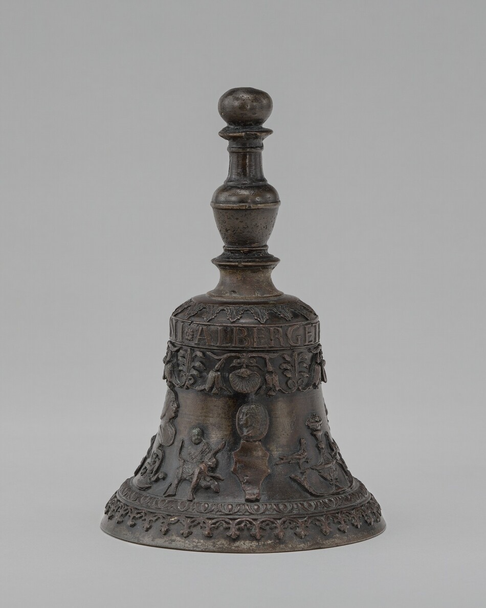 The sculpture is a decorative bell with intricate relief designs depicting scenes of figures engaged in various activities amid patterns of leaves and vines. Cast in metal, the bell has a dark bronze color with a weathered surface. The top of the bell features a rounded handle. The bell is ornamented with detailed figures and text, showcasing the craftsmanship involved in its creation.