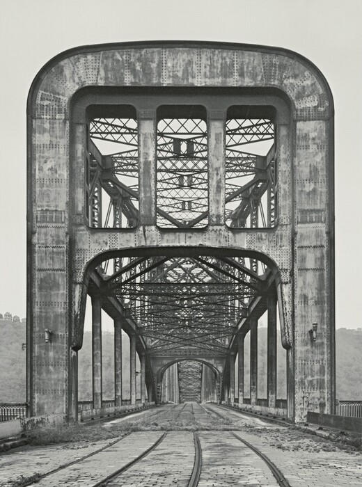 The image shows a large, industrial steel bridge with a symmetrical view. The bridge is made of metal girders and trusses creating a mesh pattern, giving depth and perspective. The pathway on the bridge is made of cobblestones leading to the central vanishing point. The sky is overcast, with minimal elements visible on the horizon and indistinct landscape features on the left side. The monochromatic color palette highlights the textures and structural details of the metal surfaces.