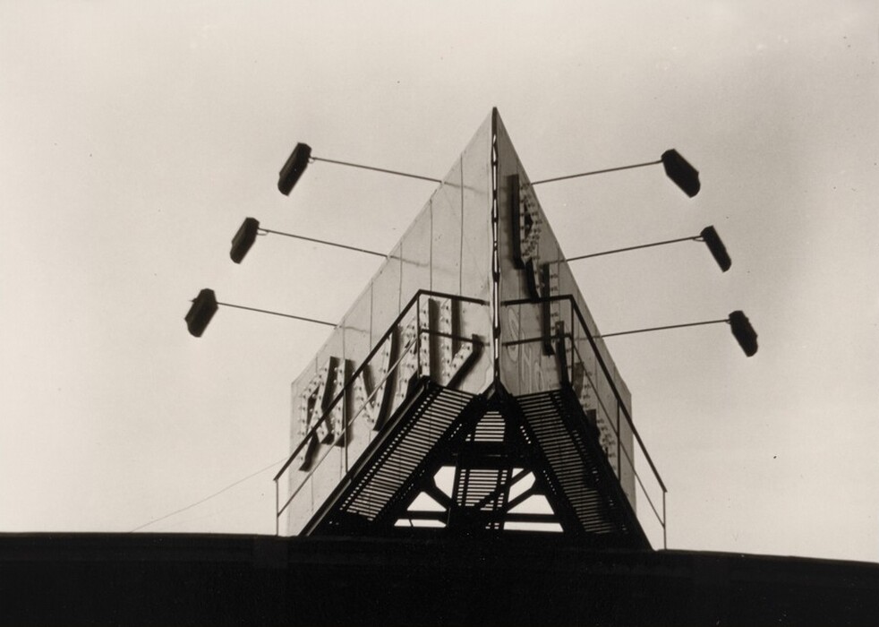 This is a photograph of a rooftop structure with a pointed sign. The image shows a large, triangular-shaped structure on top of a building, featuring a prominent metal framework with bold, angular shapes. Extending from the sides of the structure are several rod-like extensions supporting smaller rectangular elements, potentially lighting fixtures. The central triangular sign includes rows of circular elements that suggest it may have been or is illuminated. The image is composed with high contrast against a bright sky, highlighting the structure's metal and geometric design elements.