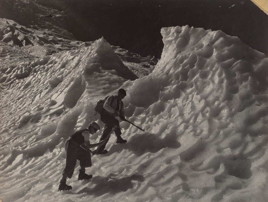 The image shows two climbers navigating a rugged, icy terrain. The scene focuses on the uneven, wave-like formations of the icy surface. The climbers are engaged in mountaineering, using crampons and ice axes. The ice shows detailed textures, carved by time and weather, giving a sense of movement and depth. The color palette is monochromatic, with shades of gray emphasizing contrasts between illuminated ice ridges and shadowed areas.
