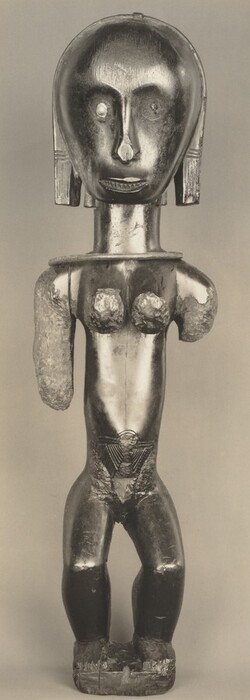 The sculpture features a figure with a large head and abstract, stylized features. The eyes are pronounced with one being a lighter spot, suggesting inlay or paint. The nose is a simple carved form, and the mouth is slightly open with visible teeth. The torso has pronounced breasts and angular, geometric patterns. The sculpture is carved from wood with a smooth, dark patina. Some areas, like the arms, show rougher textures, possibly due to the wood's natural grain or carving technique. The surface shows signs of age or wear, with cracks and patina loss at the base and chest area. The figure stands upright and is solidly connected to a base.