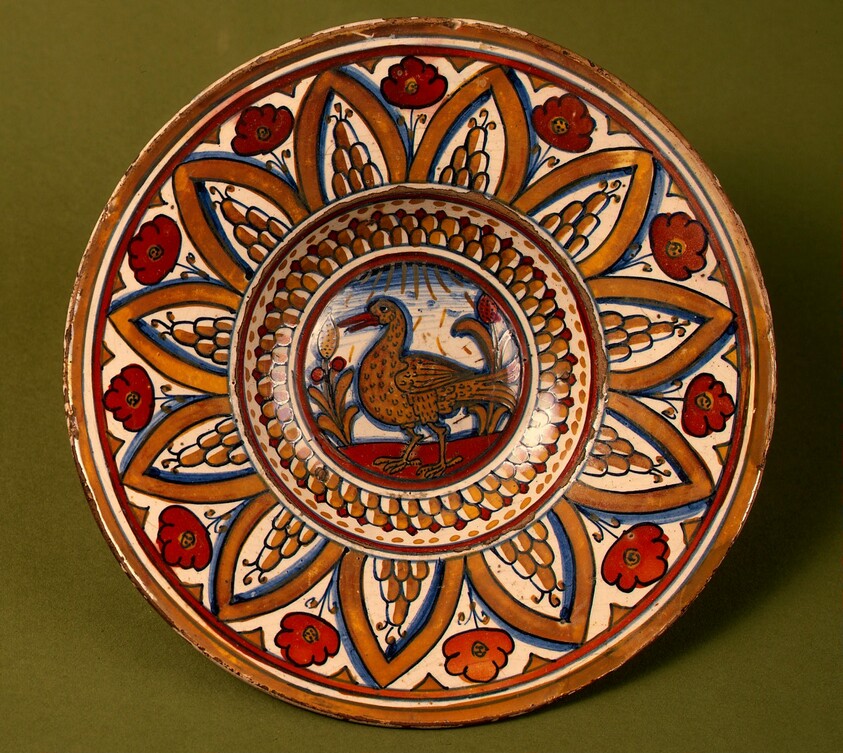 Plate with border of rounded hills within pointed arches with flowers between them; in the center, a wading bird between stylized plants