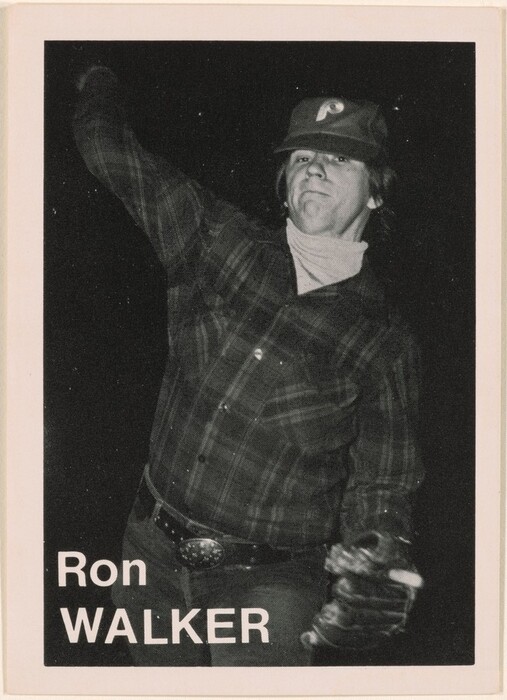 The image shows a person from mid-thigh upwards, positioned in a throwing motion with their right arm extended upwards and their left arm bent at the elbow, clad in a glove. The individual has a determined expression on their face, with light-colored hair visible beneath a baseball cap, which has a logo featuring a stylized 'P'. They are wearing a plaid shirt that is buttoned up and belted jeans, and they have a prominent belt buckle. In the background, the setting is a dark, indistinct space, suggesting that the photograph was taken in low lighting conditions, highlighting the subject. The name "Ron WALKER" is printed at the bottom left corner of the image.