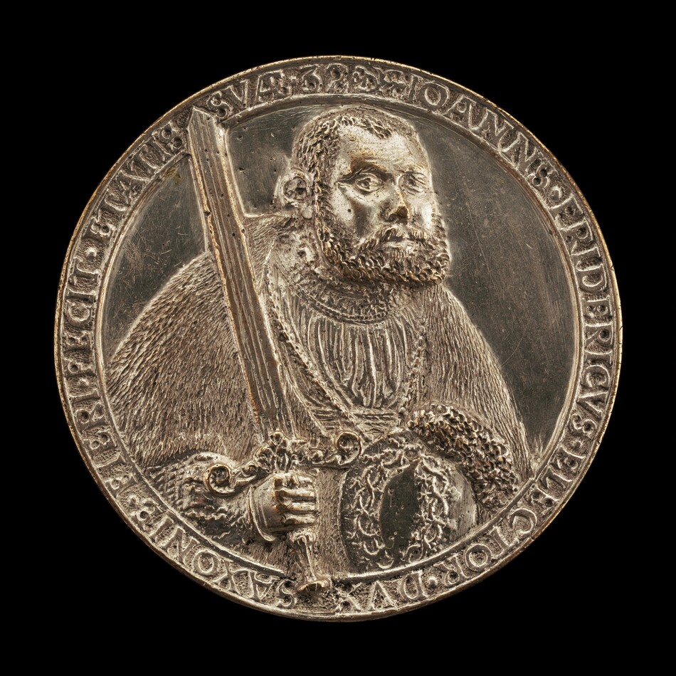 The sculpture features a man depicted in a relief on a round medal. He is shown holding a sword upright and dressed in ornate clothing with intricate patterns and textures. His beard and hair are detailed, and the material appears metallic with a silver-like sheen. Surrounding the portrait are engraved inscriptions on the border of the medal. The sculpture is relatively small, similar in size to a large coin.