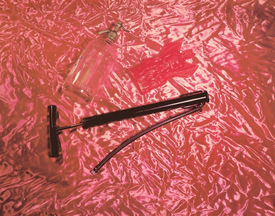 The image shows a glass bottle with a metal pump, a bicycle pump, and a small red object with an intricate design. The color palette includes vibrant pinks and reds, with a shiny, reflective fabric background.