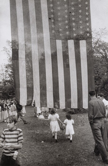 This is an image of a large American flag with people and children in the background. The flag is prominent and partially obstructs the figures behind it. The stripes and stars of the flag are visible. Below the flag is a grassy area where people are moving in various directions. Tall trees are visible on the right side of the image. Some individuals in the foreground appear to be holding balloons or objects. The image is in black-and-white.