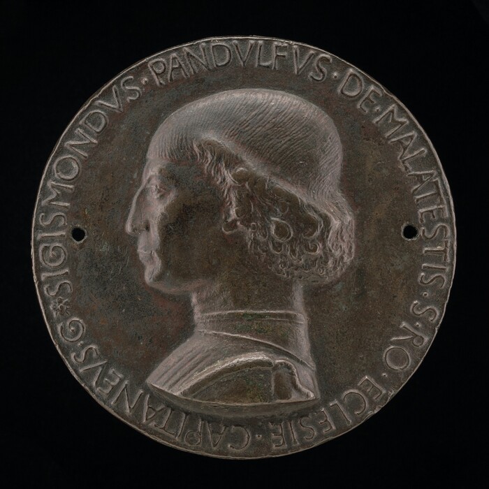 The sculpture is a circular medallion featuring a side profile of an individual. The person is looking to the left. Their hair is short and straight on the top of their head but becomes longer and curlier as it gets down to their ears. They have a flat nose and closed lips. The surface of the sculpture is embellished with inscriptions around the edge, and it is a dark brown color.
