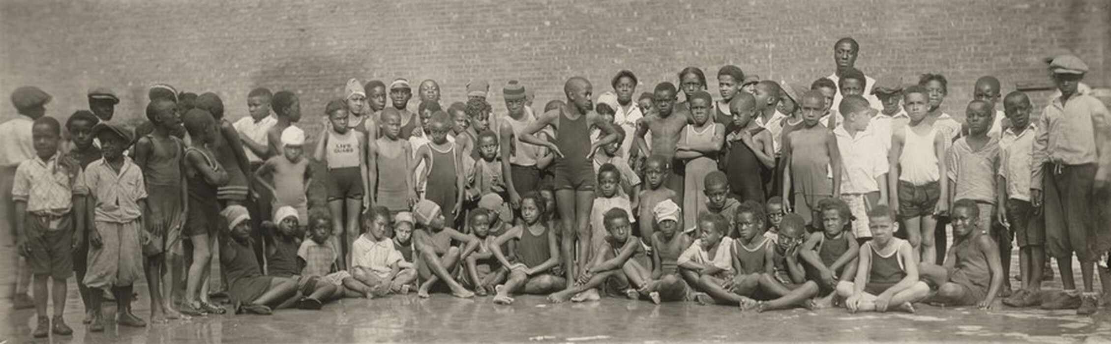 The image shows a group of children of various ages gathered in front of a brick wall. The children have diverse appearances and are dressed in casual clothing. They are either standing or sitting on the ground.