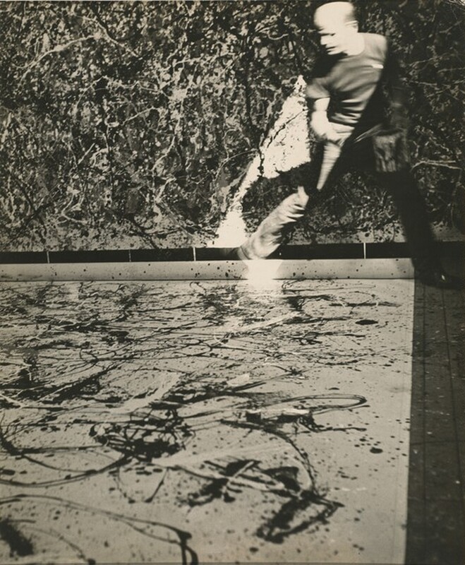 A bald, pale-skinned man holding a can of house paint stands facing us in this vertical black and white photograph. Fabric is spread across the floor, and the edge near the man is rolled up. The light-colored fabric is spattered with white and dark drips and skeins of paint. To the right, on the far side of the canvas, his legs are spread in a wide stance, one knee bent as he steps across the corner of the canvas. Having been photographed while in motion, his head and body is blurred. His head is turned to our left and his shoulders angled slightly to our right. His right side, to our left, is brightly lit by a shaft of intense light that falls from the upper right. The other side of his body is cast into deep shadow. A canvas hanging beyond him is filled with a dense web of lines and splotches in shades of black, white, and gray over a light background.