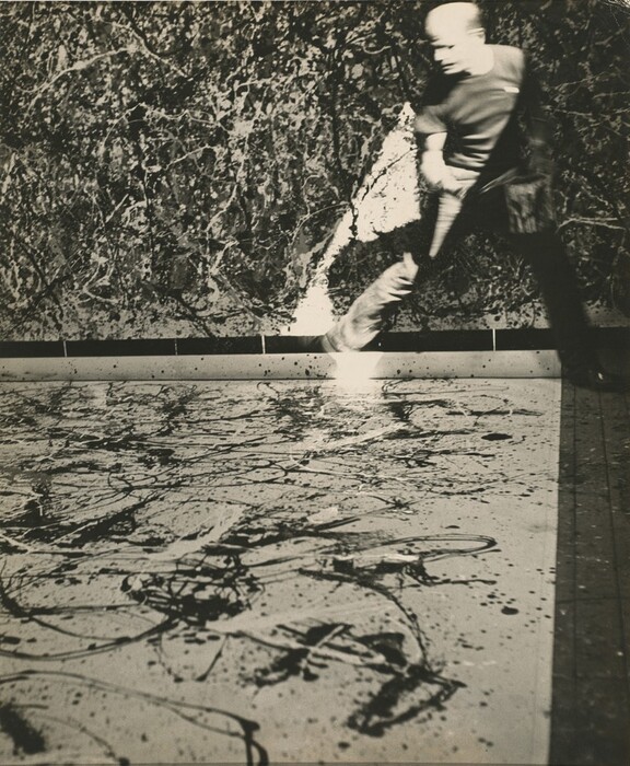 A bald, pale-skinned man holding a can of house paint stands facing us in this vertical black and white photograph. Fabric is spread across the floor, and the edge near the man is rolled up. The light-colored fabric is spattered with white and dark drips and skeins of paint. To the right, on the far side of the canvas, his legs are spread in a wide stance, one knee bent as he steps across the corner of the canvas. Having been photographed while in motion, his head and body is blurred. His head is turned to our left and his shoulders angled slightly to our right. His right side, to our left, is brightly lit by a shaft of intense light that falls from the upper right. The other side of his body is cast into deep shadow. A canvas hanging beyond him is filled with a dense web of lines and splotches in shades of black, white, and gray over a light background.
