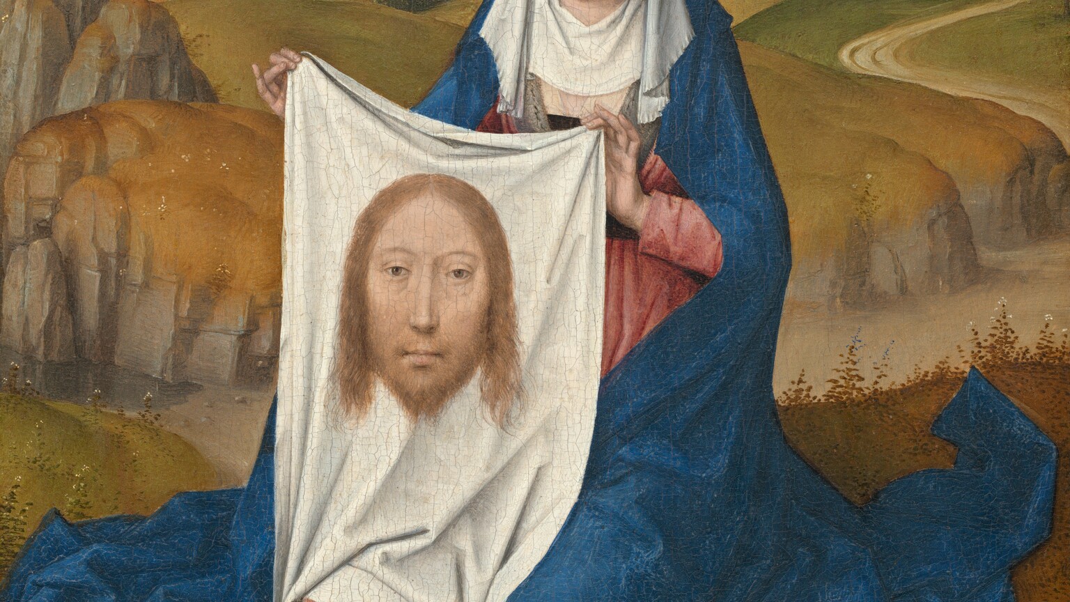 A woman sitting close to us in front of a deep landscape holds up a white cloth with the image of a man’s face on it in this vertical painting. The woman and the image on the cloth both have pale skin, hooded eyes, and long, straight noses.  The woman’s shoulders are squared toward us, and her legs are tucked under her. Her head tips slightly to our right. Her eyes are downcast, and her bow-shaped lips are parted. She wears a white, turban-like headdress with a white veil covering her neck and upper chest. Her rose-pink dress is mostly covered by a voluminous, lapis-blue robe falling from the back of the headdress to the ground, where it puddles in angular folds. On the white cloth she holds up, the man’s head has chin-length blond hair and a beard. He looks out at us with light brown eyes. The woman sits on a grassy field. A dirt path cuts through a shallow valley across the picture, not far behind the woman. More harvest-yellow and moss-green hills roll back to a town with gray stone buildings with blue roofs along the horizon, which comes three-quarters of the way up the composition. The sky above deepens from white at the horizon to azure blue across the top of the painting.