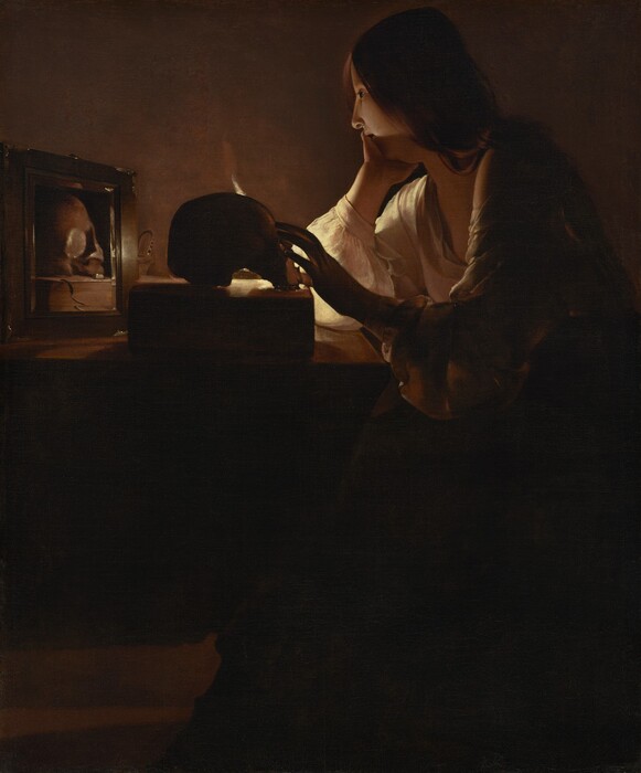 A young woman with pale skin sits at a table in a darkened room in this vertical painting. Long chestnut-brown hair drapes over her shoulder and her deep, cream-colored, long-sleeved garment is open at her neck. She rests her chin in her right hand, farther from us, as her left reaches for a skull placed on a thick book on the table in front of her. The scene is lit by a single candle mostly out of sight behind the skull. Shown in profile, she looks into a small mirror next to the skull, which reflects that object and the book.