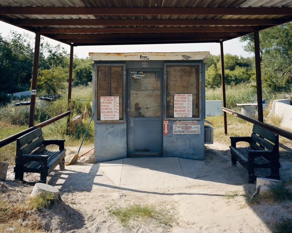 The photograph captures a small, weathered structure under a metal roof with a view slightly above eye level. The building has metal mesh windows and signs listing ferry service details, fares, and a phone number. Close to us are two black benches placed symmetrically on either side. Beyond, there is sandy ground and some vegetation leading to dense trees. The color palette is natural, with earthy tones and greenery, complemented by the subdued gray of the building and benches. Strong shadows suggest a bright, sunny day.