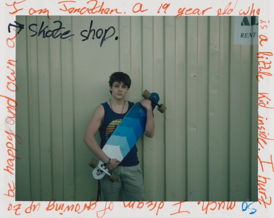 This photograph shows a person from the knees up standing in front of a corrugated metal wall. They are holding a blue and white longboard diagonally across their chest. The person has short, dark hair and pale skin. They are wearing a blue tank top with a circular design and light-colored shorts. They have a necklace around their neck and a bracelet on one wrist. The edges of the photograph are covered in handwritten text in black and orange ink on top of a white border.