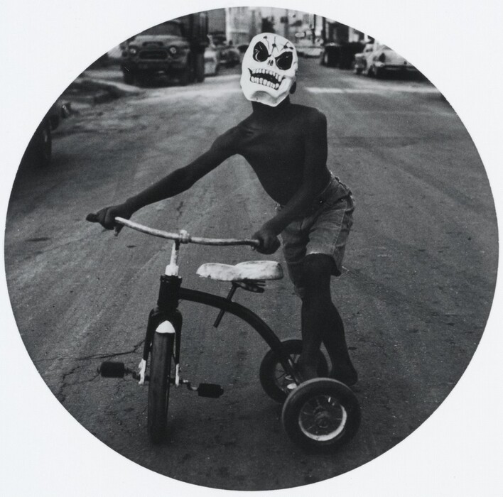 The image depicts a person riding a tricycle with a unique mask, wearing a dark top and denim shorts. The tricycle has a vintage design, and the setting is an urban street with cars parked along the sides and buildings in the background.