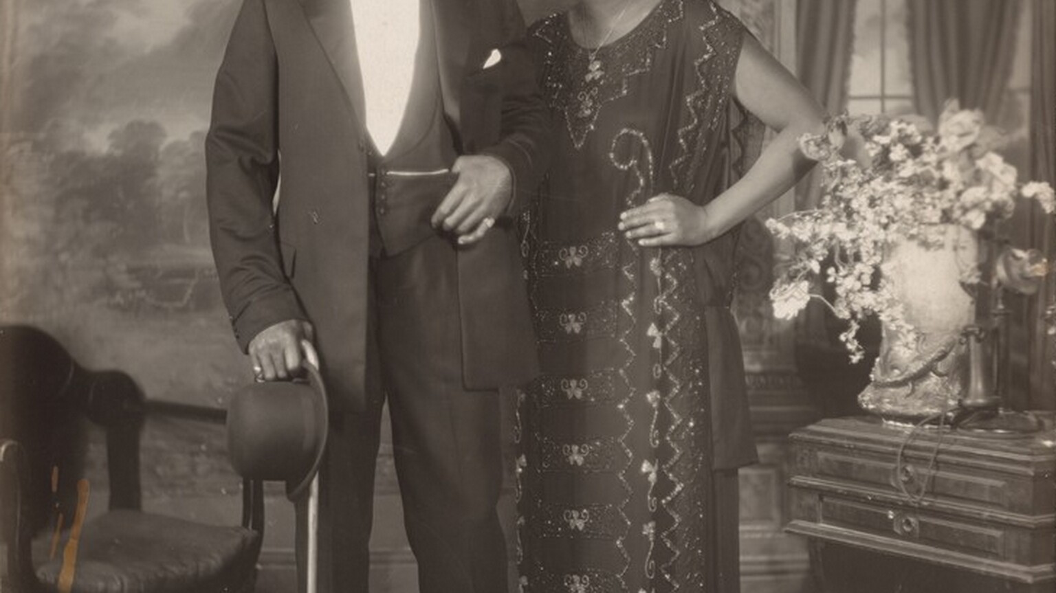 An elegantly dressed Black man and woman stand facing and looking at us, slightly smiling, in a room in this vertical photograph. The image is monochromatic like a black and white photograph but is printed in warm tones of golden and dark browns. To our left, the man has short-cropped hair and is cleanshaven. He has dark eyes, a rounded nose, and his lips are closed in a slight smile. He wears a three-piece tuxedo and holds a bowler hat and cane in his right hand, on our left. He pulls his suit jacket back to hook his other thumb in his vest pocket. He wears rings on each of his pinky fingers and a chain crosses his vest, tucked into the same pocket as his thumb. The woman stands with her left hand, on our right, on her hip and her other hand resting on the man’s shoulder. She has a delicate nose, dark eyes, and her closed lips turn up slightly at the corners. She has a cheek-length bob haircut and wears dangling earrings and a necklace with a pendant. Her ankle-length, sleeveless dress is beaded with geometric and scrolling patterns. Some of the beads and the ring she wears on the third finger of the hand on her hip catch and reflect the light. An upholstered chair sits to our left and a wood side table with an urn filled with flowers and a telephone stands to our right. The telephone has a conical earpiece hanging from a stand with a flaring mouthpiece. The backdrop behind the people has a painted or wallpapered section to our left and an arch leading to a curtained window to our right. Parts of the photograph are noticeably out of focus, particularly in the background and the flowers. The artist signed the work with white letters against the dark shadows under the seat of the chair, near the lower left corner: “VAN DER ZEE NYC 1924.”