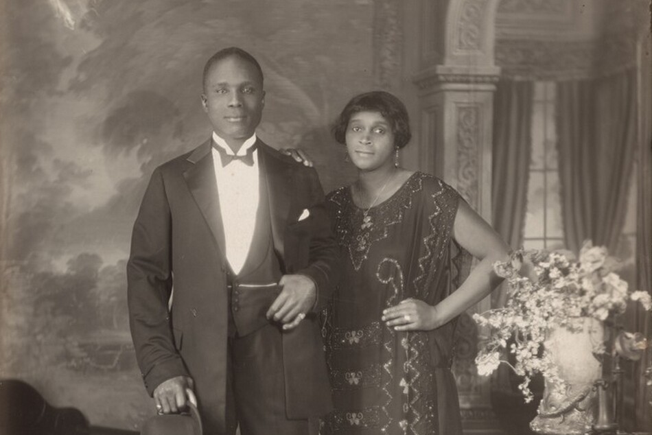 An elegantly dressed Black man and woman stand facing and looking at us, slightly smiling, in a room in this vertical photograph. The image is monochromatic like a black and white photograph but is printed in warm tones of golden and dark browns. To our left, the man has short-cropped hair and is cleanshaven. He has dark eyes, a rounded nose, and his lips are closed in a slight smile. He wears a three-piece tuxedo and holds a bowler hat and cane in his right hand, on our left. He pulls his suit jacket back to hook his other thumb in his vest pocket. He wears rings on each of his pinky fingers and a chain crosses his vest, tucked into the same pocket as his thumb. The woman stands with her left hand, on our right, on her hip and her other hand resting on the man’s shoulder. She has a delicate nose, dark eyes, and her closed lips turn up slightly at the corners. She has a cheek-length bob haircut and wears dangling earrings and a necklace with a pendant. Her ankle-length, sleeveless dress is beaded with geometric and scrolling patterns. Some of the beads and the ring she wears on the third finger of the hand on her hip catch and reflect the light. An upholstered chair sits to our left and a wood side table with an urn filled with flowers and a telephone stands to our right. The telephone has a conical earpiece hanging from a stand with a flaring mouthpiece. The backdrop behind the people has a painted or wallpapered section to our left and an arch leading to a curtained window to our right. Parts of the photograph are noticeably out of focus, particularly in the background and the flowers. The artist signed the work with white letters against the dark shadows under the seat of the chair, near the lower left corner: “VAN DER ZEE NYC 1924.”