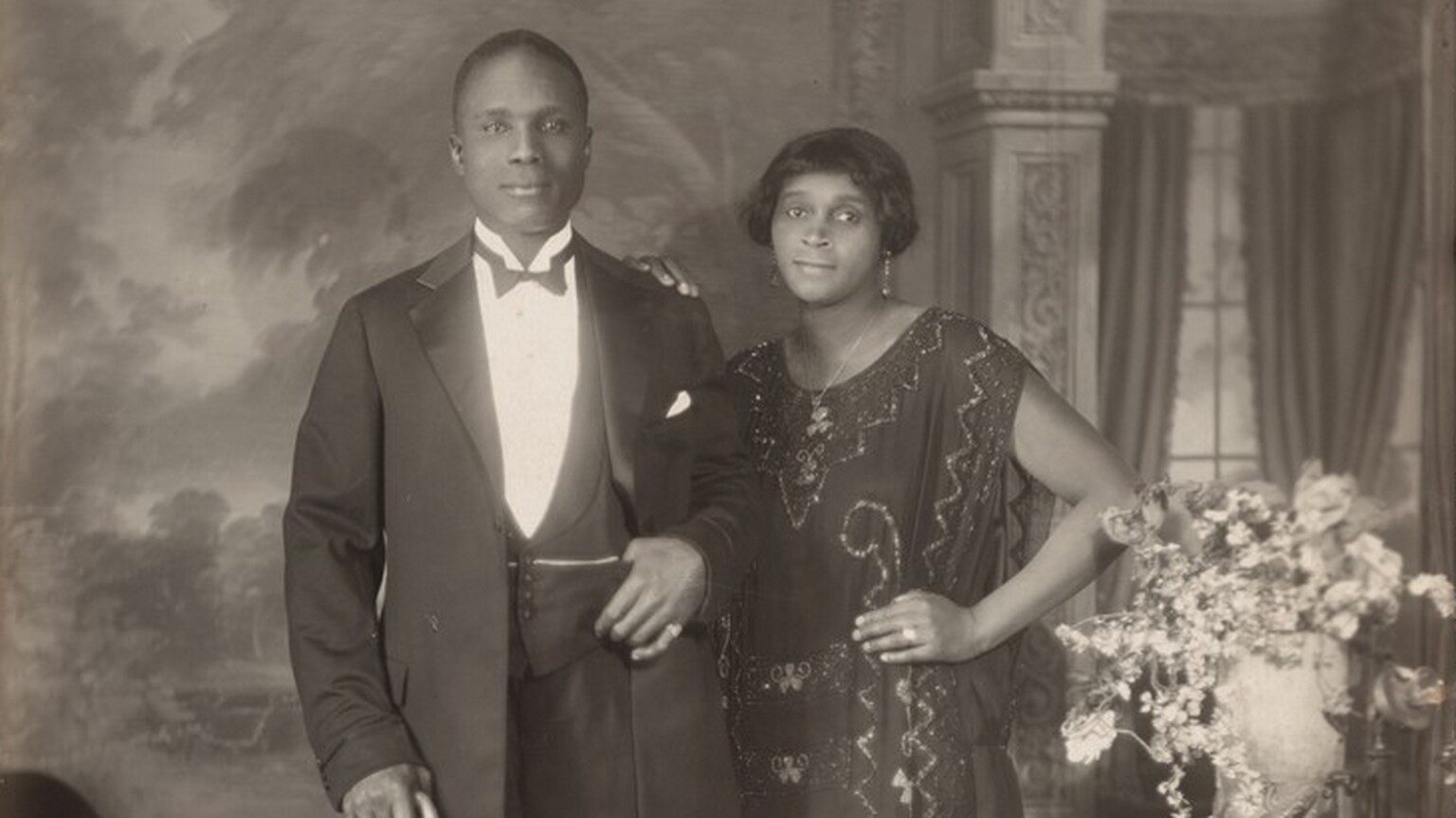 An elegantly dressed Black man and woman stand facing and looking at us, slightly smiling, in a room in this vertical photograph. The image is monochromatic like a black and white photograph but is printed in warm tones of golden and dark browns. To our left, the man has short-cropped hair and is cleanshaven. He has dark eyes, a rounded nose, and his lips are closed in a slight smile. He wears a three-piece tuxedo and holds a bowler hat and cane in his right hand, on our left. He pulls his suit jacket back to hook his other thumb in his vest pocket. He wears rings on each of his pinky fingers and a chain crosses his vest, tucked into the same pocket as his thumb. The woman stands with her left hand, on our right, on her hip and her other hand resting on the man’s shoulder. She has a delicate nose, dark eyes, and her closed lips turn up slightly at the corners. She has a cheek-length bob haircut and wears dangling earrings and a necklace with a pendant. Her ankle-length, sleeveless dress is beaded with geometric and scrolling patterns. Some of the beads and the ring she wears on the third finger of the hand on her hip catch and reflect the light. An upholstered chair sits to our left and a wood side table with an urn filled with flowers and a telephone stands to our right. The telephone has a conical earpiece hanging from a stand with a flaring mouthpiece. The backdrop behind the people has a painted or wallpapered section to our left and an arch leading to a curtained window to our right. Parts of the photograph are noticeably out of focus, particularly in the background and the flowers. The artist signed the work with white letters against the dark shadows under the seat of the chair, near the lower left corner: “VAN DER ZEE NYC 1924.”