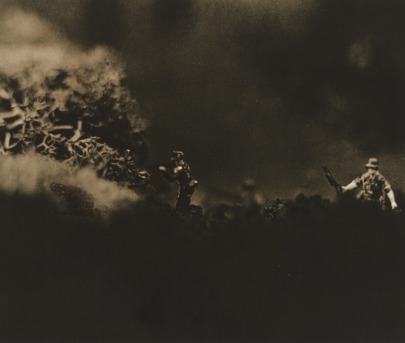 This abstract image features dark and blurred shapes dominating the scene, with two small, detailed figures in the foreground standing out against the hazy background. The composition creates an impression of a miniature landscape, where the figures resemble toy soldiers positioned as if encountering a rough terrain. The large, textured mass on the left side of the image could be compared to a rock or dense foliage, while the muted lighting enhances the mysterious, undefined quality of the scene. Overall, the arrangement of shapes and lines draws a connection to a dramatic diorama or a model battlefield.