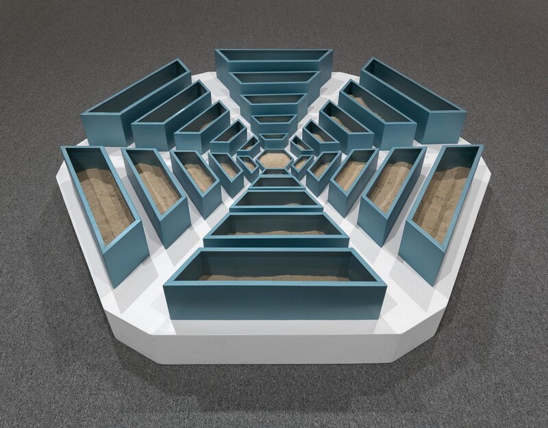 We look slightly down at a hexagonal, abstract sculpture made with teal-blue, wooden bins radiating out in six arms from a smaller hexagonal bin in the center. Each of the six arms tapers from wider and taller bins at the perimeter to narrower, shorter bins near the center. Light brown sand partially fills the bins. The bins sit on a white platform, the outlines of which follow the six sides of the snowflake-like arrangement of bins. The platform sits on battleship-gray carpet.