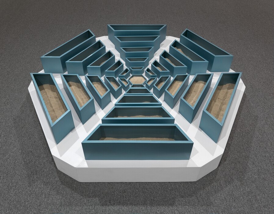 We look slightly down at a hexagonal, abstract sculpture made with teal-blue, wooden bins radiating out in six arms from a smaller hexagonal bin in the center. Each of the six arms tapers from wider and taller bins at the perimeter to narrower, shorter bins near the center. Light brown sand partially fills the bins. The bins sit on a white platform, the outlines of which follow the six sides of the snowflake-like arrangement of bins. The platform sits on battleship-gray carpet.