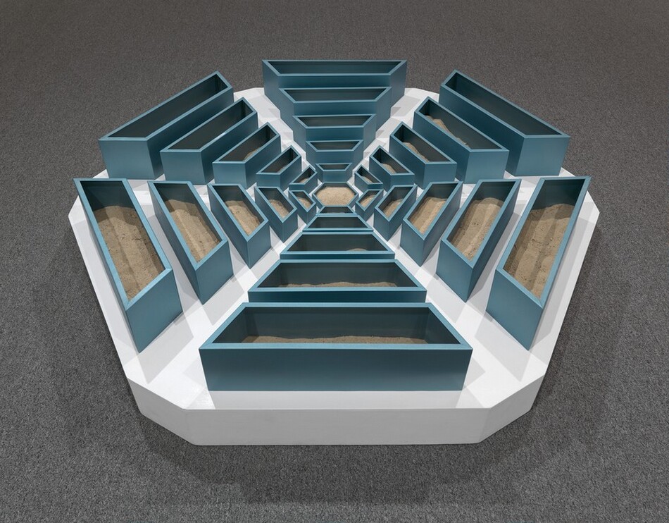 We look slightly down at a hexagonal, abstract sculpture made with teal-blue, wooden bins radiating out in six arms from a smaller hexagonal bin in the center. Each of the six arms tapers from wider and taller bins at the perimeter to narrower, shorter bins near the center. Light brown sand partially fills the bins. The bins sit on a white platform, the outlines of which follow the six sides of the snowflake-like arrangement of bins. The platform sits on battleship-gray carpet.