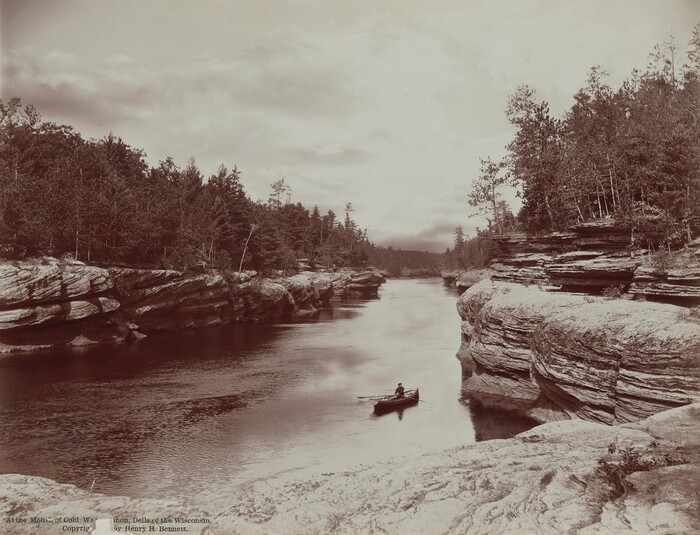 From the flat top of striated, sheer rocks, we look down onto a person rowing a canoe toward us in a river in this horizontal black and white photograph. The rocky banks funnel the river, which comes toward us, like a canal. Evergreen trees are spiky silhouettes against a cloudy, pale sky. Printed text at the bottom left of the photograph reads, “At the Mouth of Cold Water Canyon, Dells of the Wisconsin. Copyright by Henry H. Bennett.”