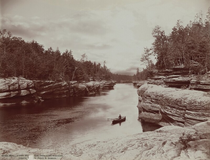 From the flat top of striated, sheer rocks, we look down onto a person rowing a canoe toward us in a river in this horizontal black and white photograph. The rocky banks funnel the river, which comes toward us, like a canal. Evergreen trees are spiky silhouettes against a cloudy, pale sky. Printed text at the bottom left of the photograph reads, “At the Mouth of Cold Water Canyon, Dells of the Wisconsin. Copyright by Henry H. Bennett.”