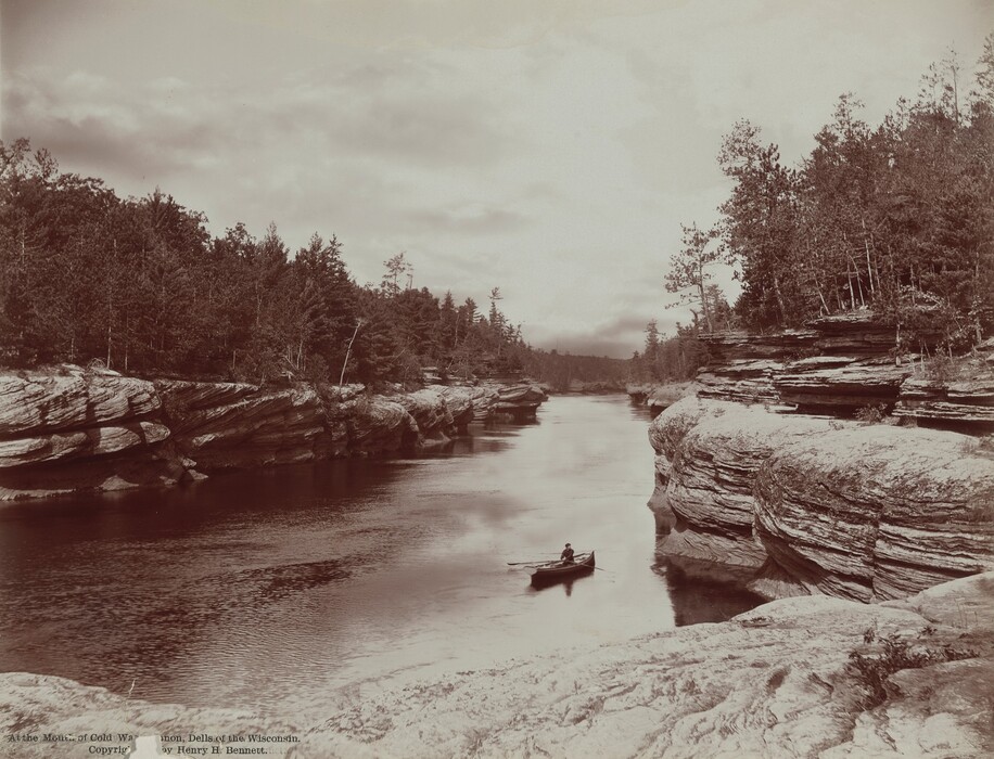 From the flat top of striated, sheer rocks, we look down onto a person rowing a canoe toward us in a river in this horizontal black and white photograph. The rocky banks funnel the river, which comes toward us, like a canal. Evergreen trees are spiky silhouettes against a cloudy, pale sky. Printed text at the bottom left of the photograph reads, “At the Mouth of Cold Water Canyon, Dells of the Wisconsin. Copyright by Henry H. Bennett.”