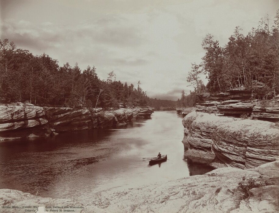 From the flat top of striated, sheer rocks, we look down onto a person rowing a canoe toward us in a river in this horizontal black and white photograph. The rocky banks funnel the river, which comes toward us, like a canal. Evergreen trees are spiky silhouettes against a cloudy, pale sky. Printed text at the bottom left of the photograph reads, “At the Mouth of Cold Water Canyon, Dells of the Wisconsin. Copyright by Henry H. Bennett.”