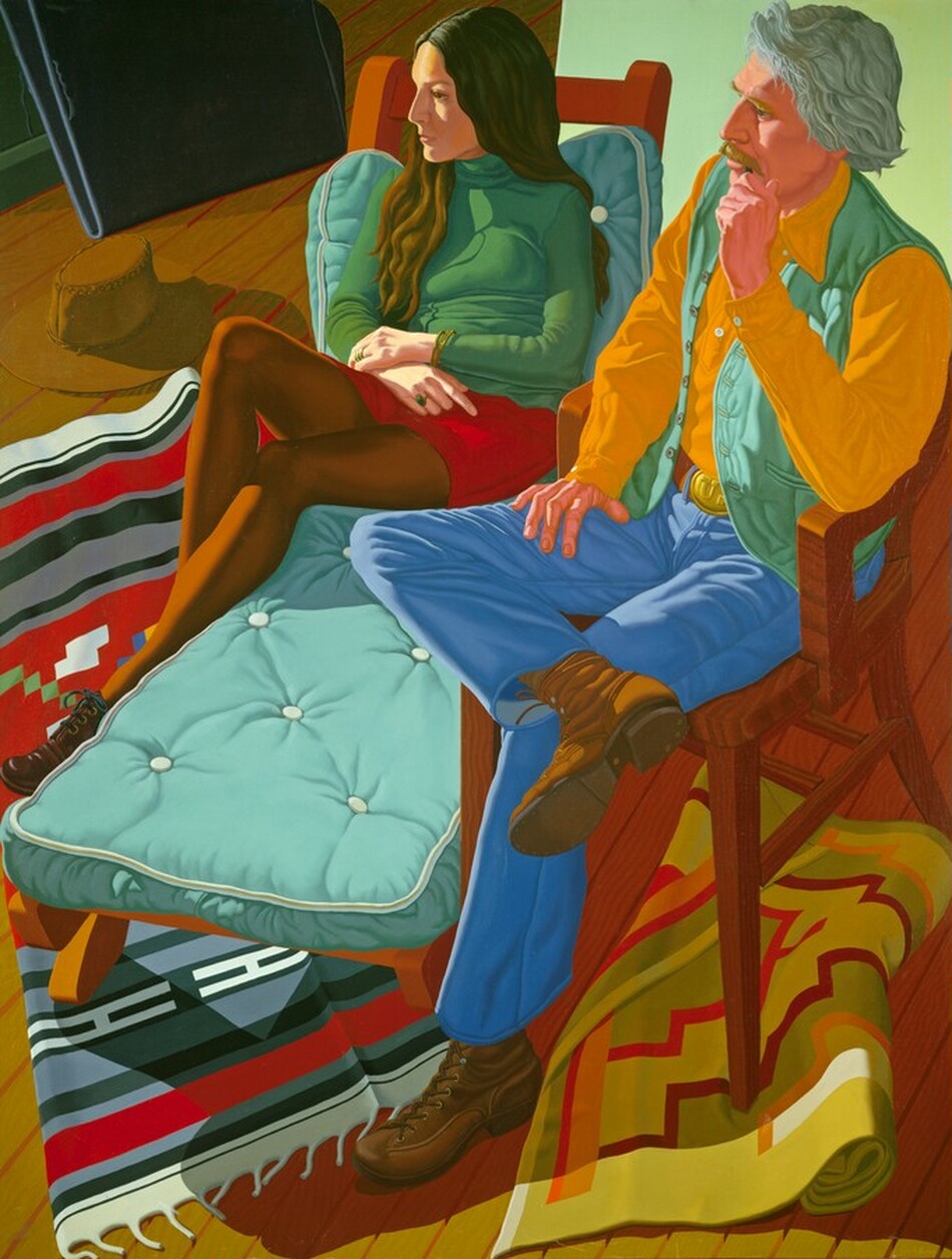 This painting shows two people sitting, looking towards the left, with their full bodies visible. The person on the left is a woman with light skin and long, dark brown hair. She is seated on a cushioned teal chaise lounge with her legs crossed. She is wearing a green turtleneck sweater and a short red skirt, with dark tights and black shoes. She has a bracelet on her wrist and rings on her fingers. Next to her, on the left, a brown cowboy hat rests near the edge of a colorful, southwestern-patterned rug. The man on the right is seated on a wooden chair next to the chaise lounge. He has light skin with gray hair and a mustache, and appears contemplative, touching his chin with one hand. He wears a yellow shirt, a light green vest, and blue pants, with a large gold belt buckle and brown shoes. More geometric patterned rugs lie beneath the man's chair. The room has wooden floors in warm, earthy tones, and a minty green wall.