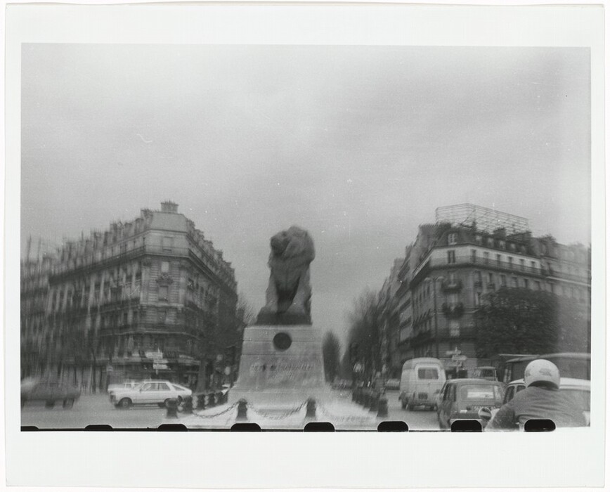 The image is a black and white photograph taken from a street-level perspective. The camera is pointed toward a large stone monument in the center of the frame, depicting a lion statue on a pedestal. The horizon is not distinctly visible, as the buildings and monument rise above the horizon line. The buildings are positioned to the left and right of the monument, forming a symmetrical urban setting leading toward the statue. The architecture consists of multi-story classic European buildings, and vehicles, including old-style cars and a motorcycle, are visible close to us, blending into the street scene. The image has a slightly blurred, out-of-focus quality, adding to the vintage feel. The color palette is limited to shades of gray and white, characteristic of the monochrome film photography style.