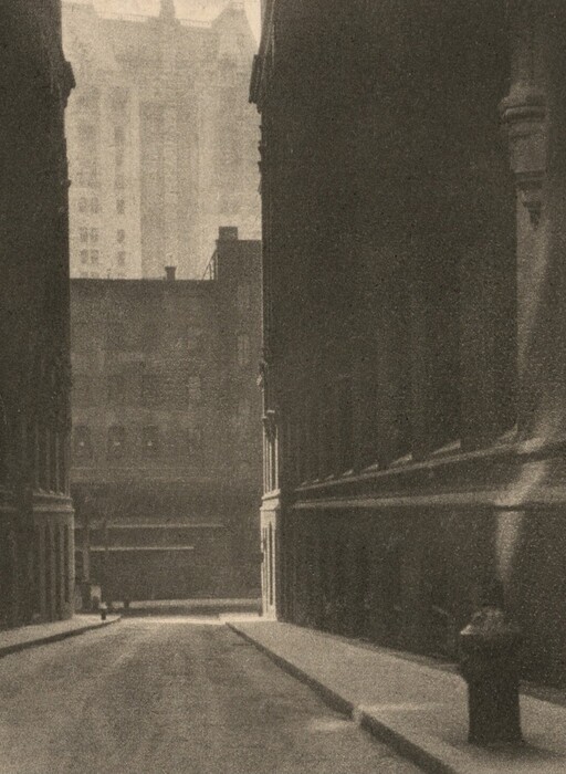 The image depicts a narrow urban alleyway flanked by tall buildings on both sides. The alley stretches into the distance, with a more illuminated area visible far ahead. The buildings create a tunnel-like effect, guiding the viewer's gaze. The photo has a grainy texture, giving it an atmospheric quality. The sepia tones create a vintage look, emphasizing shadow and light contrast. In the distance, faintly visible structures add depth to the urban environment.
