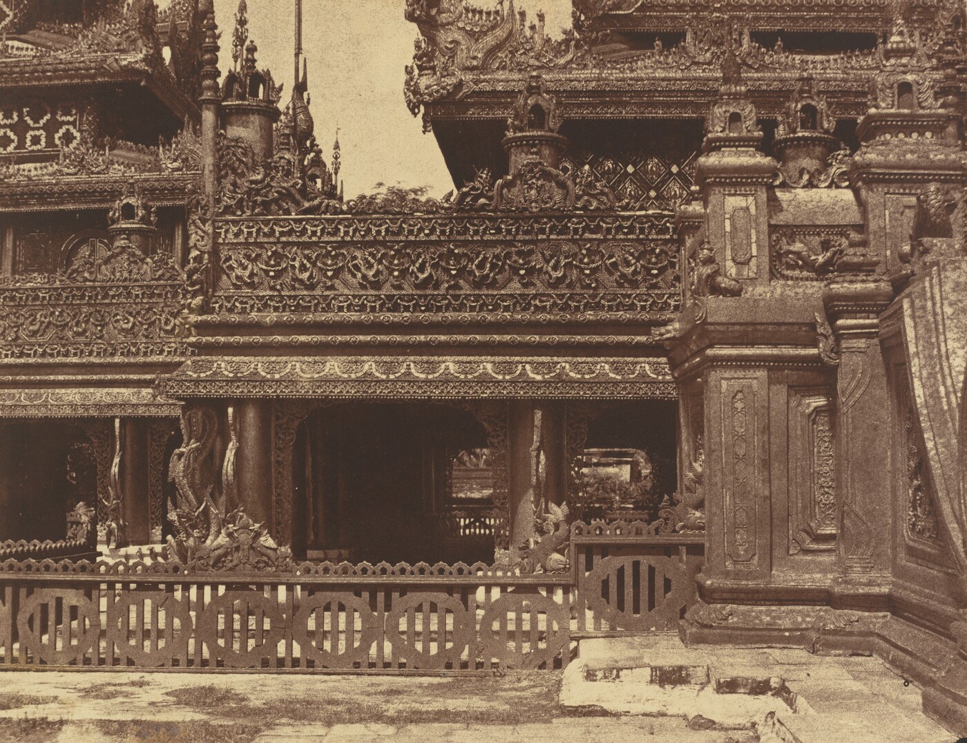 This is a photograph of an intricately carved building. The image shows the architectural details of the structure, featuring elaborate wooden carvings and ornamental designs. The building is adorned with lattice works, motifs, animals, and mythical creatures, showcasing detailed craftsmanship. A decorative railing is visible at the lower part of the building, while the upper sections display sculptural elements, spires, and decorative panels. The design reflects traditional Southeast Asian architecture, highlighted in great detail in the photograph.