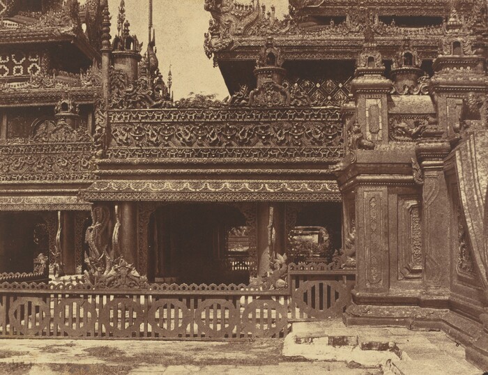This is a photograph of an intricately carved building. The image shows the architectural details of the structure, featuring elaborate wooden carvings and ornamental designs. The building is adorned with lattice works, motifs, animals, and mythical creatures, showcasing detailed craftsmanship. A decorative railing is visible at the lower part of the building, while the upper sections display sculptural elements, spires, and decorative panels. The design reflects traditional Southeast Asian architecture, highlighted in great detail in the photograph.