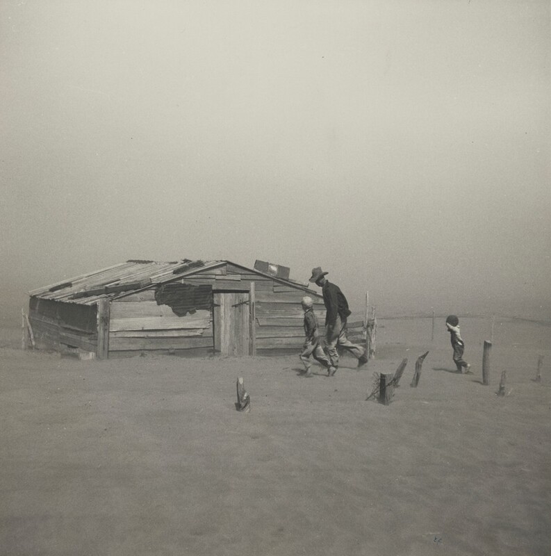 A man and two children dash for a dilapidated, wooden structure in a flat, deserted landscape in this horizontal photograph. The dusty sky and sandy ground blend together in a haze. The man is tall and wears a dark coat and hat. His head tipped down so the brim protects his face, and he leans forward. Next to him is a child wearing a white cap. A few steps behind them, to our right, is another child with his or her hands in the air. The building is a low, A-frame structure made of broken, horizontal wood planks. The gently sloping tin roof is held down by what seems to be larger pieces of wood. The door is in the center of the side we see, and is constructed of vertical planks of wood fastened by dark metal bits. To the left of the door a large hole appears to have been patched in the side of the structure.