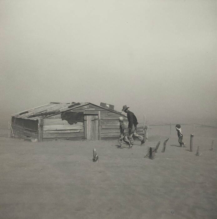 A man and two children dash for a dilapidated, wooden structure in a flat, deserted landscape in this horizontal photograph. The dusty sky and sandy ground blend together in a haze. The man is tall and wears a dark coat and hat. His head tipped down so the brim protects his face, and he leans forward. Next to him is a child wearing a white cap. A few steps behind them, to our right, is another child with his or her hands in the air. The building is a low, A-frame structure made of broken, horizontal wood planks. The gently sloping tin roof is held down by what seems to be larger pieces of wood. The door is in the center of the side we see, and is constructed of vertical planks of wood fastened by dark metal bits. To the left of the door a large hole appears to have been patched in the side of the structure.