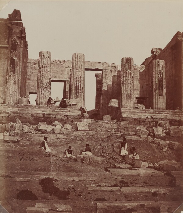The image shows an upward view of an ancient architectural structure with large columns and ruins. The surface is uneven with scattered stones and debris. Several people, dressed in traditional clothing, are among the remnants of the structure. The color palette is sepia-toned, giving the image a vintage look with tones of brown and tan.