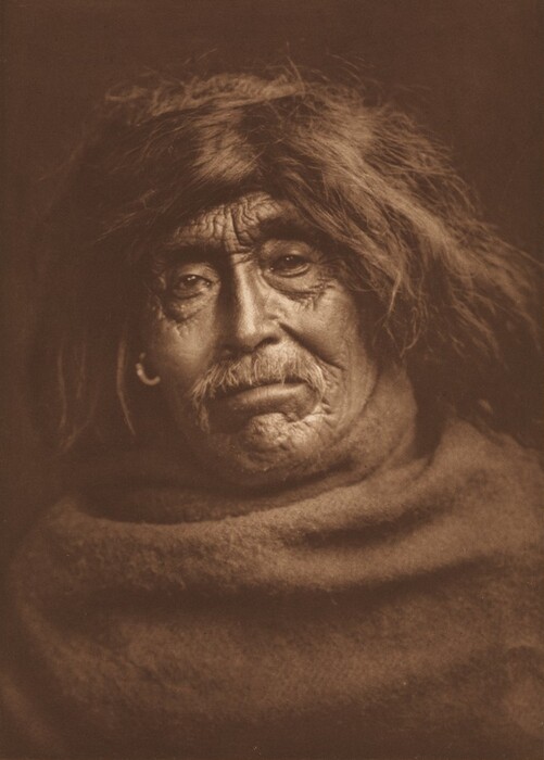 The image shows the upper body and face of an elderly person in a seated position. The person's facial features are prominent, with noticeably weathered skin and deep wrinkles, especially around the eyes and forehead. They have a mustache, and their hair is long, thick, and tousled, reaching around their face and partially obscuring the forehead. The hair appears dark with signs of graying. They are draped in a textured fabric that covers their shoulders. They are wearing a simple earring in one ear. The background is a plain, dark backdrop.