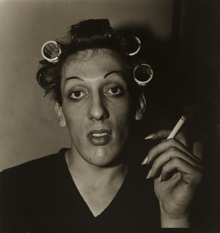 This nearly square black and white photograph shows the head and shoulders of a person in front of a plain wall. Wearing a dark V-necked shirt, the person has an oval-shaped face and dark hair rolled in curlers. Finely plucked dark eyebrows frame large, luminous dark eyes, and the full lips are slightly open. A lit cigarette is held aloft in the person’s long-nailed left hand, on our right. Light comes from the left and deepens into shadow across the right half of the photograph.