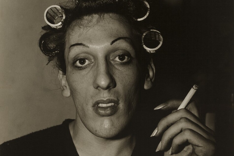 This nearly square black and white photograph shows the head and shoulders of a person in front of a plain wall. Wearing a dark V-necked shirt, the person has an oval-shaped face and dark hair rolled in curlers. Finely plucked dark eyebrows frame large, luminous dark eyes, and the full lips are slightly open. A lit cigarette is held aloft in the person’s long-nailed left hand, on our right. Light comes from the left and deepens into shadow across the right half of the photograph.