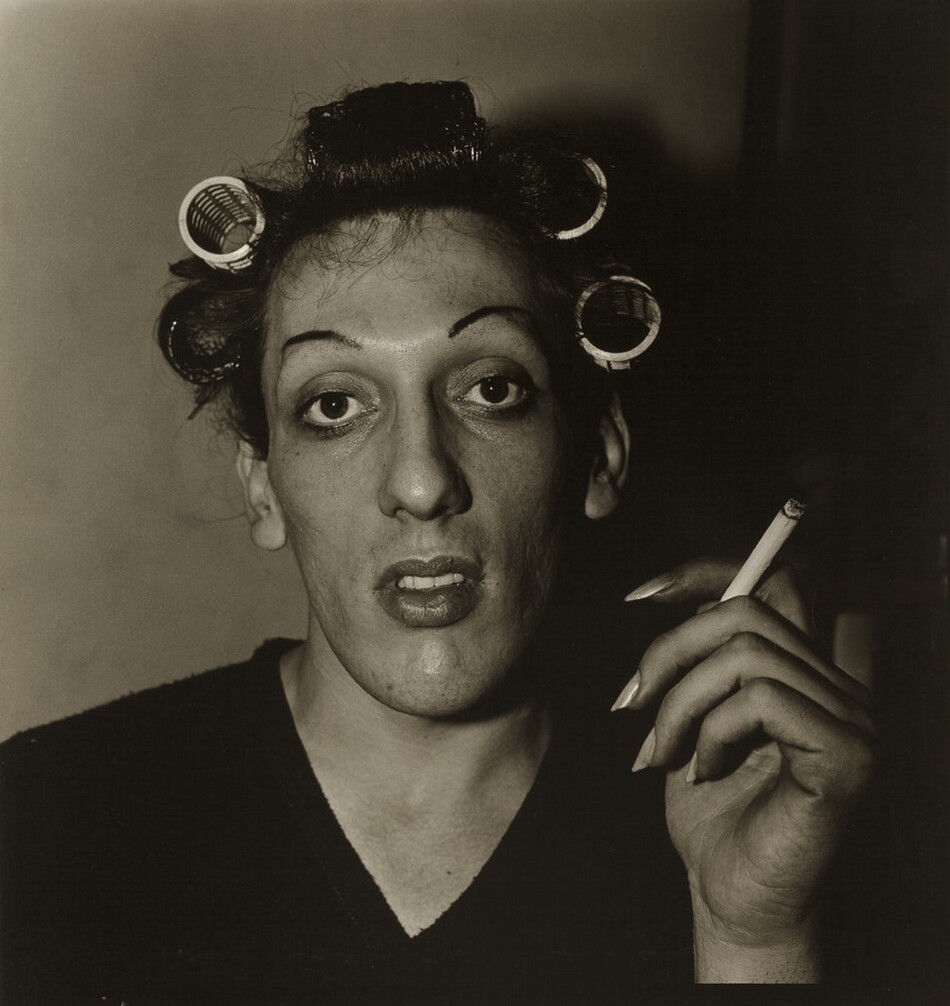 This nearly square black and white photograph shows the head and shoulders of a person in front of a plain wall. Wearing a dark V-necked shirt, the person has an oval-shaped face and dark hair rolled in curlers. Finely plucked dark eyebrows frame large, luminous dark eyes, and the full lips are slightly open. A lit cigarette is held aloft in the person’s long-nailed left hand, on our right. Light comes from the left and deepens into shadow across the right half of the photograph.